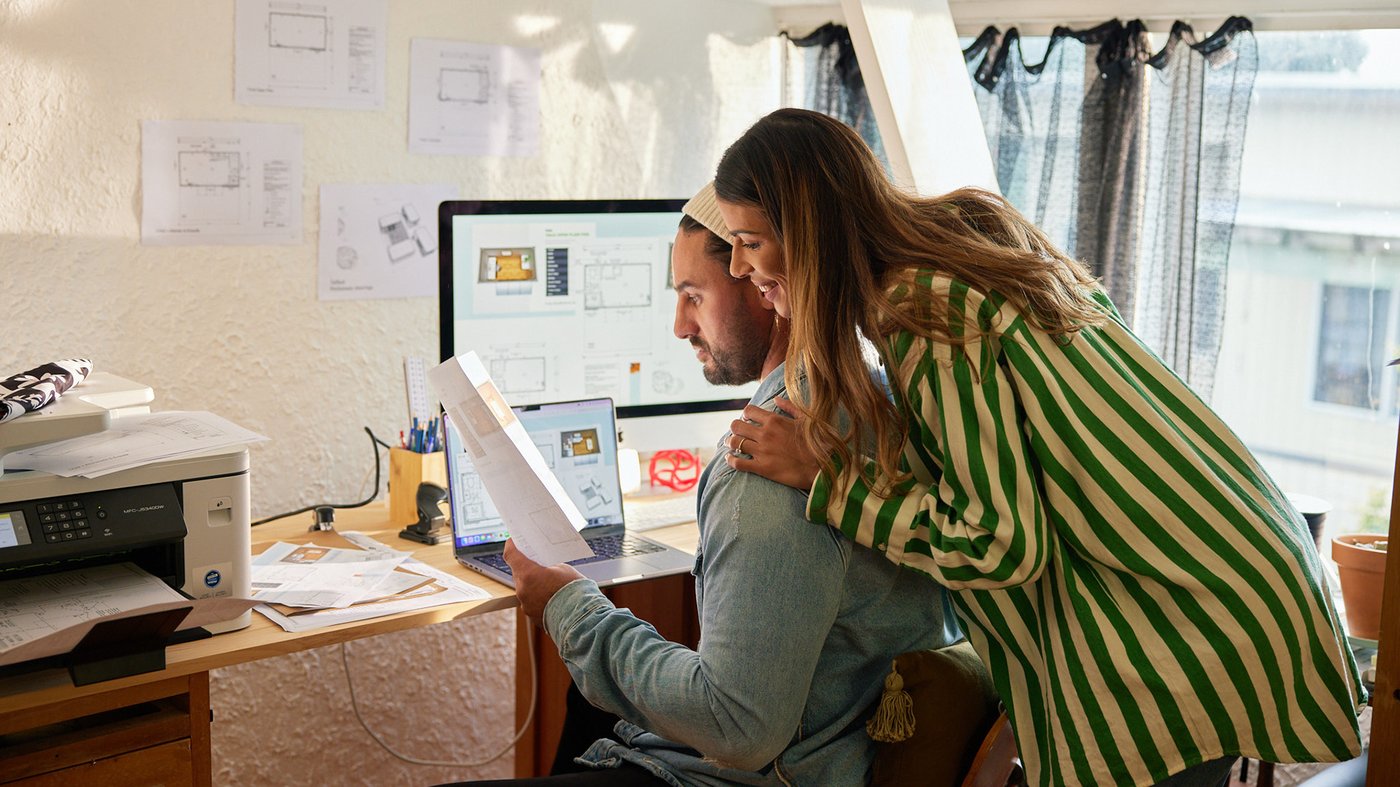 A couple reviews printed plans at a home office desk, with design drawings on the wall and a printer beside them.