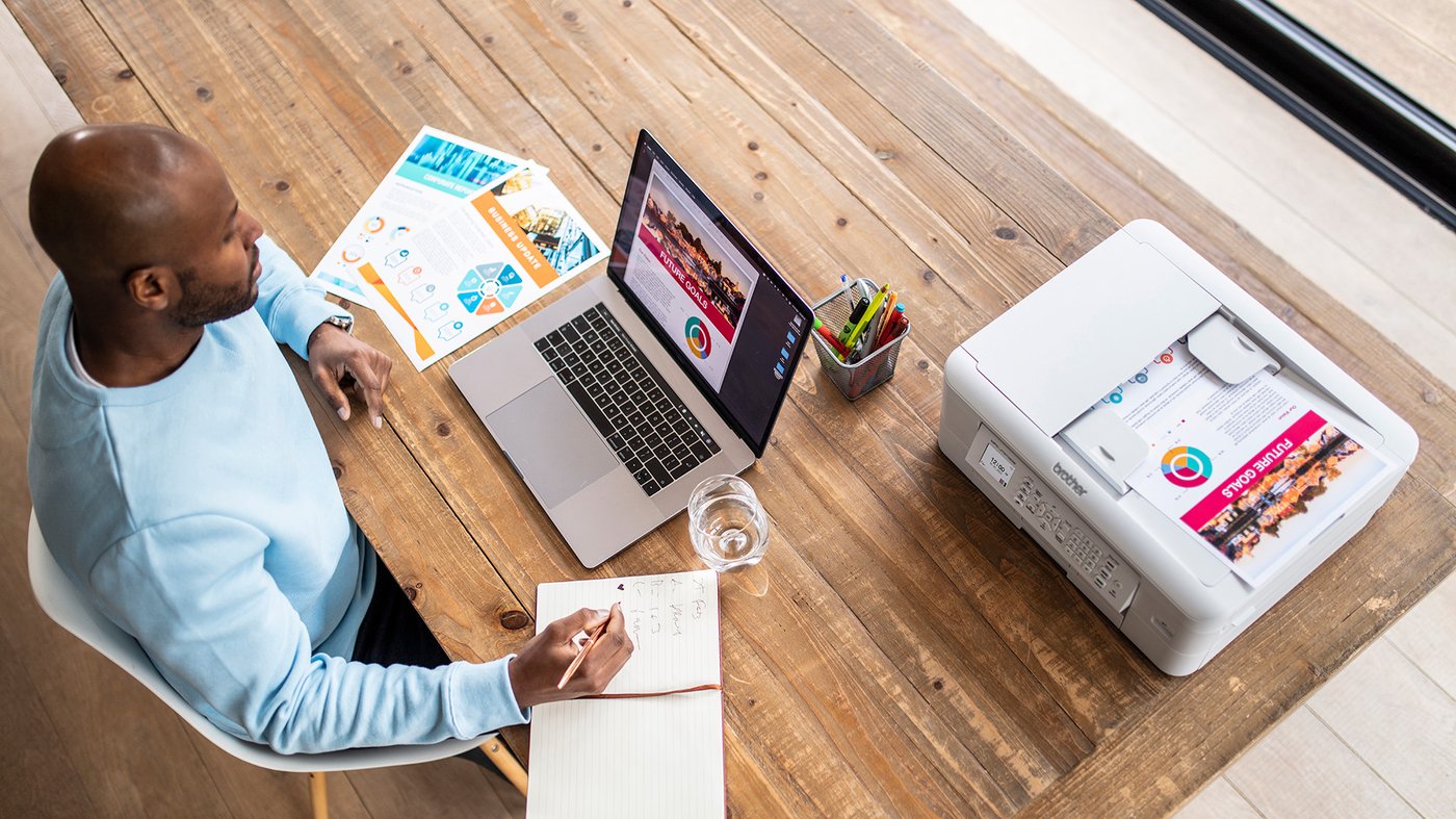 A man sits at a wooden table working on a laptop, with colourful printed documents beside him and a Brother inkjet printer producing a full colour page.