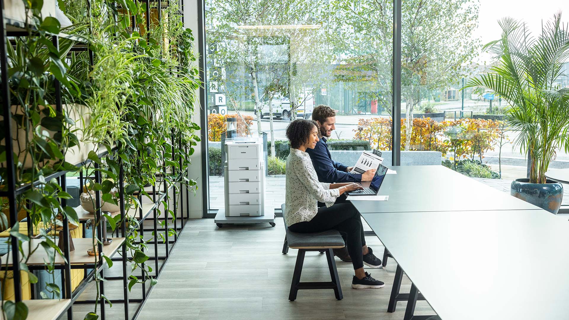 Two people working on laptops in a modern office with abundant indoor plants and a Brother multifunction printer in the background, highlighting sustainable workplace printing.